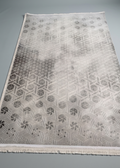 A rectangular area rug with a geometric pattern, shown flat on a neutral gray background. The rug features a gradient design that transitions from a light cream-white at the top to a darker gray at the bottom, with a fringed edge.