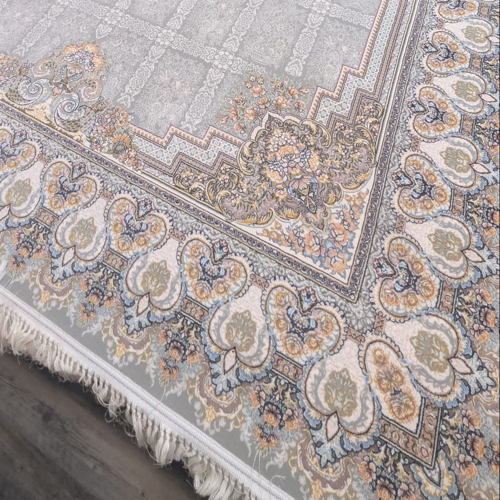 A large rectangular area rug with a traditional Persian-style pattern, featuring a central medallion, ornate borders, and fringe on the short ends. The rug has a light gray base with beige, blue, and rust accents.