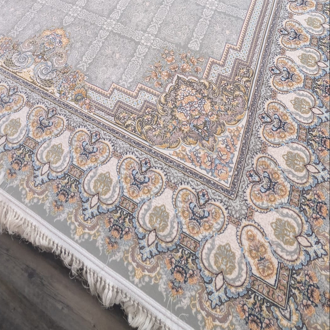 A large rectangular area rug with a traditional Persian-style pattern, featuring a central medallion, ornate borders, and fringe on the short ends. The rug has a light gray base with beige, blue, and rust accents.