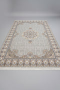 A rectangular area rug with a traditional Persian-style floral pattern, laid out on a wooden floor in what appears to be a showroom or warehouse. The rug features a light beige and gray base with cream, gold, and muted blue accents in its intricate medallion and border designs.