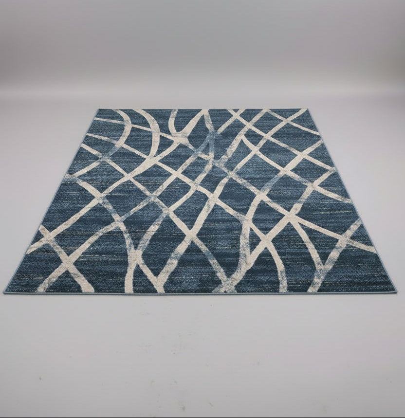 A rectangular area rug with a blue and off-white geometric lattice pattern, displayed on a table in a showroom with other rugs in the background.