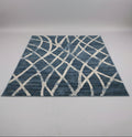 A rectangular area rug with a blue and off-white geometric lattice pattern, displayed on a table in a showroom with other rugs in the background.