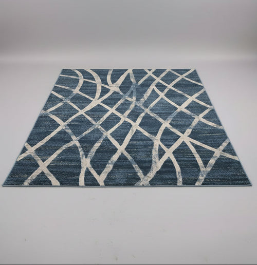 A rectangular area rug with a blue and off-white geometric lattice pattern, displayed on a table in a showroom with other rugs in the background.
