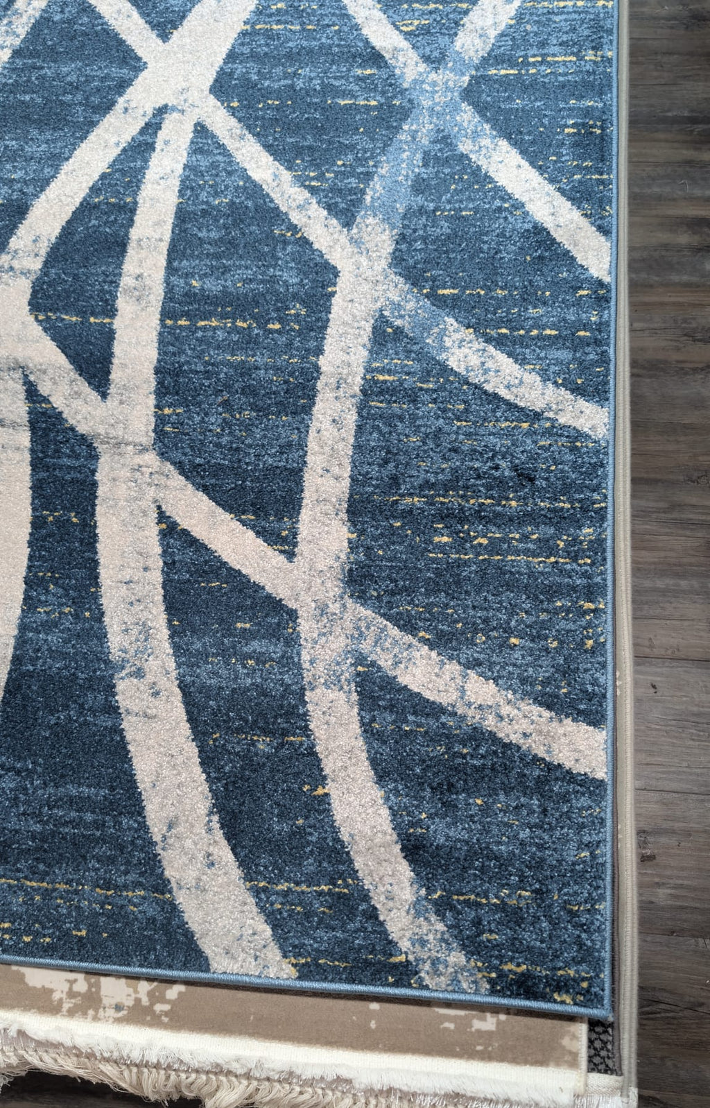 A close-up of a blue area rug with a distressed abstract pattern of cream-colored curved lines and a fringed edge, shown on a dark wood floor.