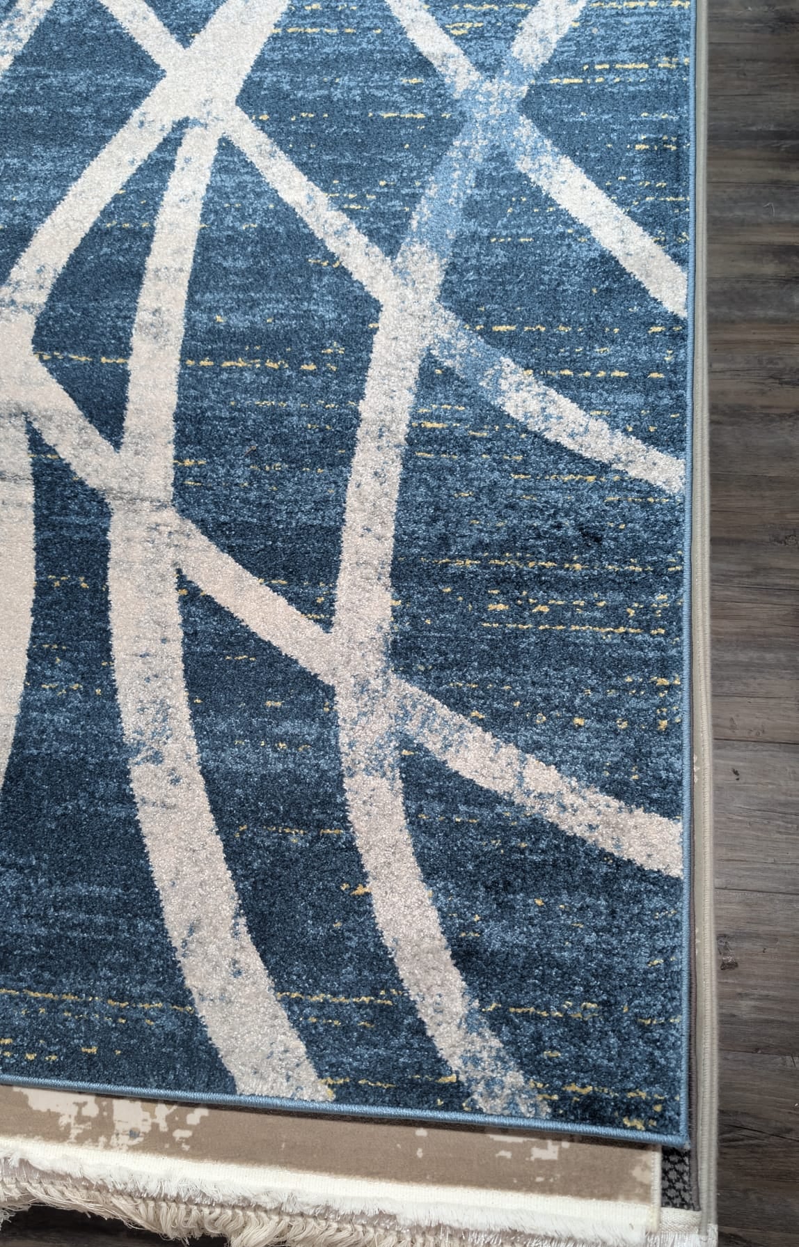 A close-up of a blue area rug with a distressed abstract pattern of cream-colored curved lines and a fringed edge, shown on a dark wood floor.