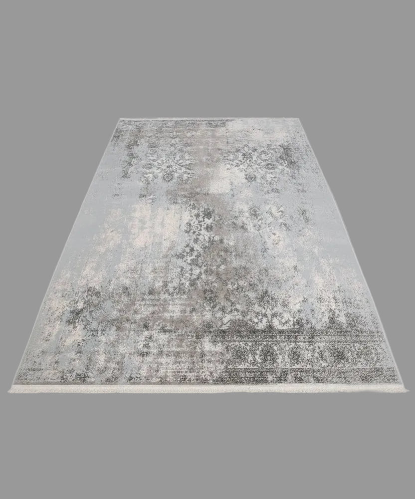 A large rectangular area rug with a distressed traditional pattern in shades of gray, beige, and off-white, laid out on a wooden floor. A person's feet in white sneakers are visible at the bottom edge of the frame.